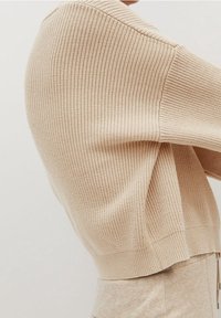 Béžový žebrovaný pletený svetr s dlouhými rukávy, který má volný střih a zaoblený lem. Měkký materiál s jemným pleteným vzorem.