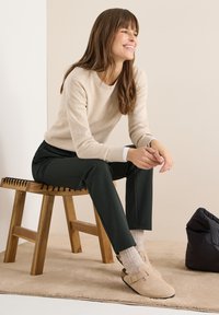 Kvinna i beige tröja, mörka byxor och beigea slip-on skor sittande på en träbänk på en beige matta bredvid en svart väska.