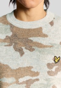 Hellblaues und sandfarbenes gestricktes Sweatshirt mit einem Camouflage-Muster. Hat einen runden Halsausschnitt und ein kleines gelbes besticktes Logo auf der Brust.