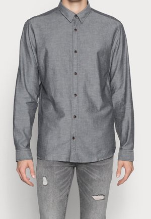 Mand iført langærmet grå skjorte med knapper, kombineret med lysegrå, revnede jeans, stående foran en ensfarvet baggrund.