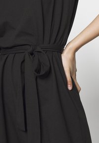 Czarna sukienka z wiązanym pasem, miękkiego materiału i bocznymi kieszeniami. Krój ma luźny fason i delikatnie opada na ciało.