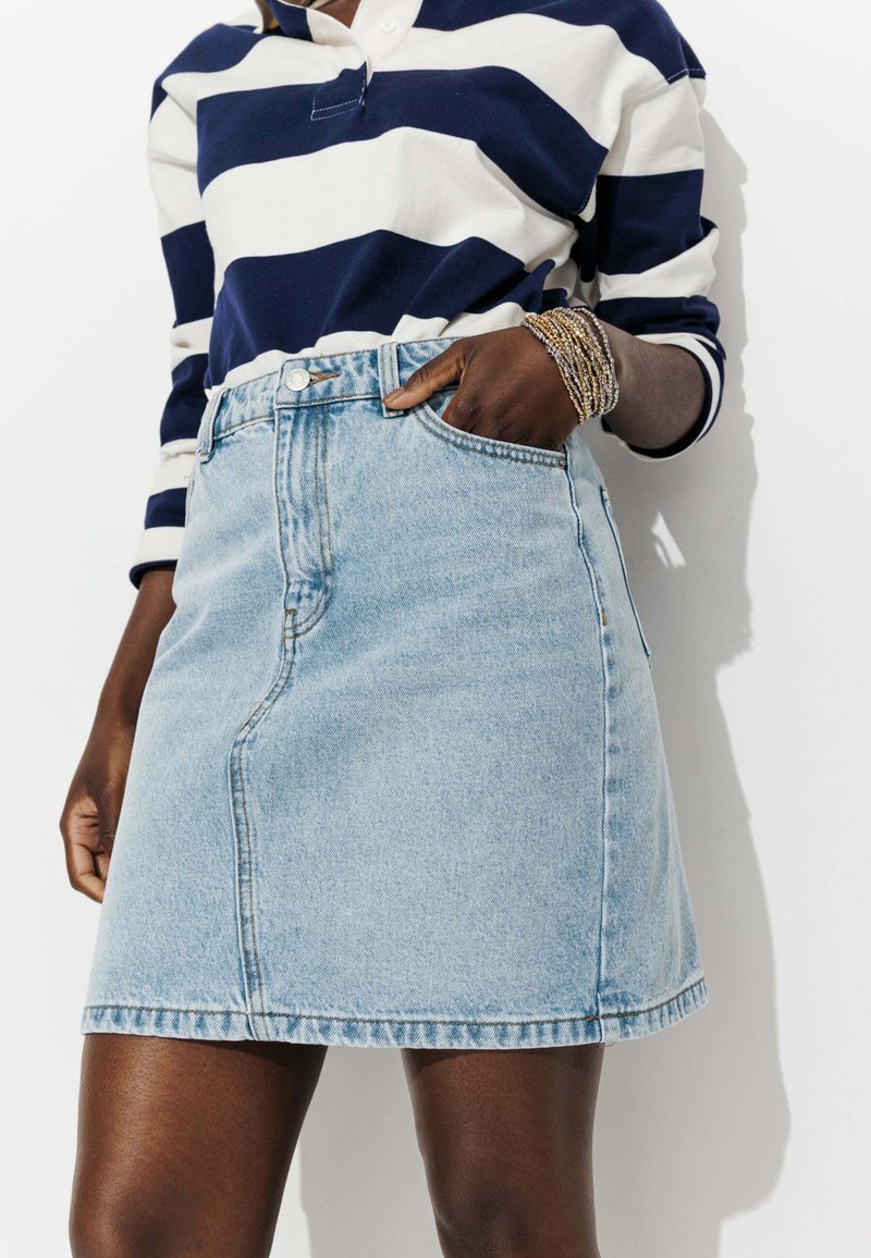 Jupe en denim délavé avec une taille ajustée, des détails de couture subtils et des poches. Associée à une chemise à manches longues rayée bleu marine et blanche.