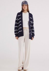 Námořnický pruhovaný kardigan přes bílou crop top, doplněný světle béžovými širokými kalhotami a bílými botami s červenými a modrými akcenty.