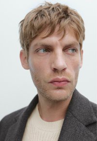 Primo piano di un uomo con capelli biondi corti e disordinati, leggera barba, occhi verdi che guarda di lato, indossando un maglione crema e un cappotto grigio scuro.