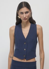 Gilet blu navy con scollatura a V profonda, cinque bottoni e tasche laterali. Vestibilità aderente e tessuto liscio con orlo corto.
