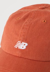 Καπέλο μπέιζμπολ σε σκουριά απόχρωση με λευκό κεντημένο λογότυπο New Balance στην μπροστινή πλευρά, που δείχνει την υφή του υφάσματος και τις λεπτομέρειες της ραφής.