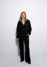 Schwarze zip-up Kapuzenjacke mit besticktem Text, kombiniert mit passenden Hosen. Weicher Stoff, lockere Passform und lange Ärmel. Einfaches Design.