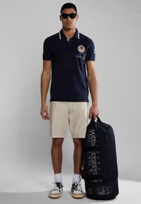 Námořnické polo tričko s logem, béžové kraťasy, bílé ponožky a tenisky. Drží velkou černou cestovní tašku s potištěným brandingem.