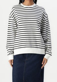 Sudadera a rayas en algodón blanco y negro con cuello redondo. Presenta mangas largas, dobladillo acanalado y ajuste relajado. Combinada con una falda vaquera oscura.