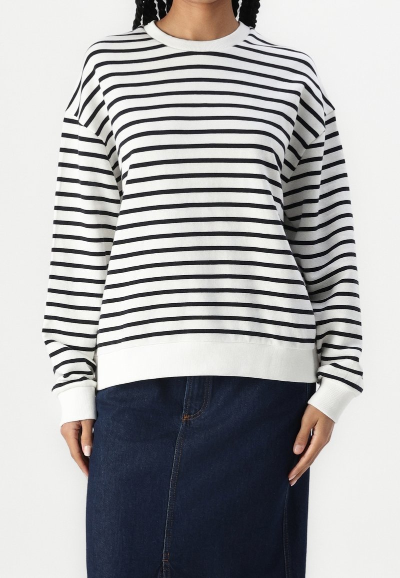 Sudadera a rayas en algodón blanco y negro con cuello redondo. Presenta mangas largas, dobladillo acanalado y ajuste relajado. Combinada con una falda vaquera oscura.