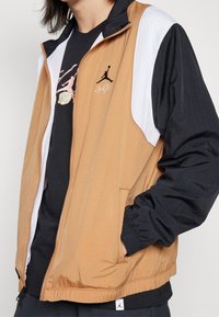 Kevyt vetoketjullinen takki ruskeana, mustana ja valkoisena. Ominaisuuksiin kuuluu joustavat hihansuut, sivutaskut ja musta Jumpman-logo rinnassa.