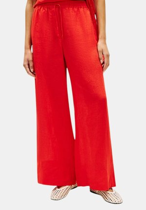 Røde bukser med vide ben og snøre i taljen, båret med hvide vævede slip-on sko, vist fra talje til fødder.