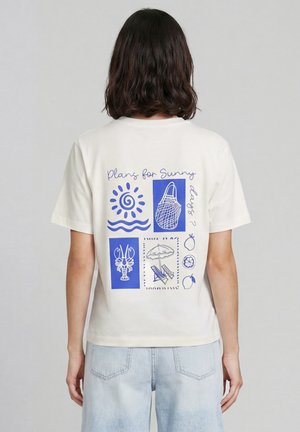 Personne portant un t-shirt blanc avec des graphismes bleus sur le thème de l'été et le texte « Plans pour les journées ensoleillées ? » dans le dos, associé à un jean bleu clair.