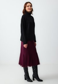 Pull noir à col roulé haut, associé à une jupe bordeaux avec des accents plissés. Le modèle porte des bottes noires à talons montantes.