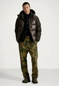 Man wearing shiny dark brown puffer jacket, black buttoned sweater, camouflage cargo pants, and dark sneakers standing with hands in pockets.