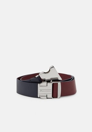 Tweedelige leren riem in marineblauw en bordeauxrood met een zilverkleurige metalen klikgesp, opgerold op een witte achtergrond.