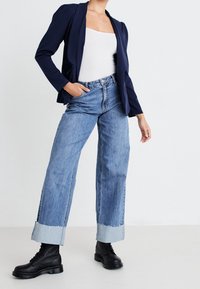 Námornícky blazer přes bílý top bez rukávů, doplněný o světle modré džíny s vysokým pasem a roztrhanými manžetami, a černé šněrovací boty.