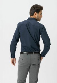 Mand set fra bagfra iført en navy blue langærmet skjorte stoppet ned i grå bukser med et sort bælte, mod en hvid baggrund.