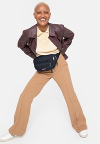 Veste en cuir marron, polo beige côtelé, sac banane marine, pantalon large beige, baskets blanches, mannequin souriant posant contre un fond blanc.