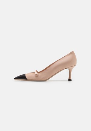 Zapato de mujer beige con puntera puntiaguda, capuchón negro en la punta, pequeño detalle de lazo y tacón delgado de altura media sobre fondo blanco.