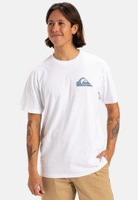 Camiseta de algodón blanca con un pequeño gráfico de olas multicolores en el lado izquierdo del pecho. Mangas cortas y un diseño de corte relajado.