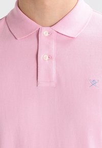 Polo rose pâle, tissu texturé, patte de boutonnage à deux boutons, petit logo brodé en bleu, col classique, design épuré.