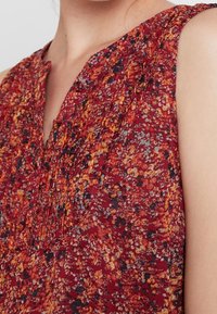 Blusa senza maniche con motivo floreale rosso, caratterizzata da una texture plissettata, un collo a V e dettagli con bottoni. Tessuto morbido e leggero.