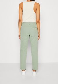 Kobieta stoi tyłem, ubrana w jasne zielone spodnie o prostych nogawkach, białą bezrękawnik i białe trampki na jednolitym tle.