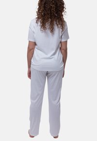 Nainen, jolla on kihara hius, seisoo paljain jaloin selkä kameraan, päällään valkoinen lyhythihainen paita ja löysät valkoiset housut tasaisella taustalla.