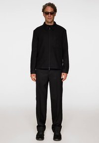 Svart jacka med dragkedja och krage, kombinerad med svarta vida byxor och sneakers. Jackan har en bröstficka och ett texturerat tyg.