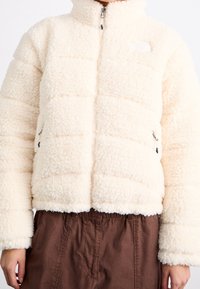 Kurtka polarowa w kolorze kremowym z pikowanym wzorem, wysoka stójka, zamek błyskawiczny z przodu i boczne kieszenie. Miękka tekstura, krótki kroj.