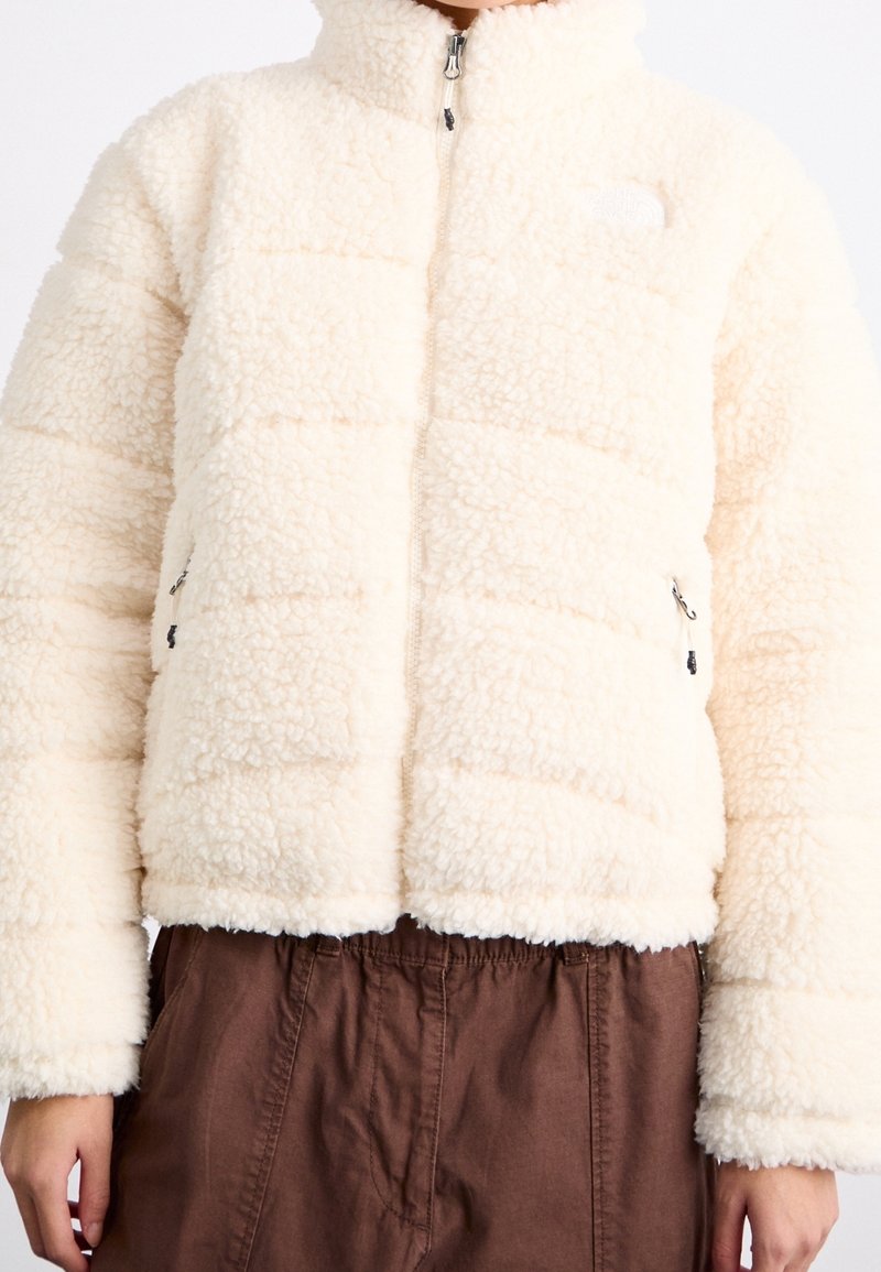 Kurtka polarowa w kolorze kremowym z pikowanym wzorem, wysoka stójka, zamek błyskawiczny z przodu i boczne kieszenie. Miękka tekstura, krótki kroj.