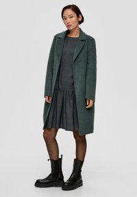 Waldgrüner Wollmantel mit eingekerbten Revers, über einem karierten Kleid. Schwarze Ankle Boots mit klobigen Sohlen. Schlichter Hintergrund.