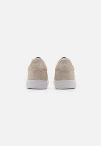 Béžové tenisky s hladkým syntetickým svrškem, bílou gumovou podešví a reliéfním logem na patě. Jednoduchý a minimalistický design.