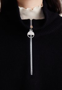 Schwarzer gerippter Strickpullover mit hohem Kragen und silbernem Reißverschluss, ausgestattet mit einem runden metallischen Zip-Ziehen und einer kontrastierenden weißen Unterlage.