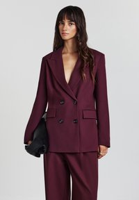 Blazer doppiopetto in un profondo bordeaux con ampi risvolti, tessuto testurizzato e bottoni neri. Abbinato a pantaloni coordinati e una pochette nera.
