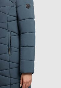 Padded vonkajšia bunda v tmavomodrej farbe s prešívaným vzorom, obsahujúca nášivku s logom, zipsový vrecko a hladkú textúru na rukáve.