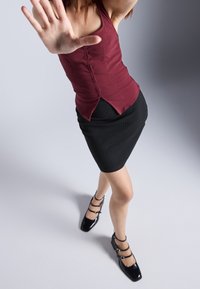 Haut ajusté bordeaux avec des coutures verticales. Jupe crayon noire à rayures. Chaussures noires en vernis avec des accents de boucle. La pose du mannequin obscurcit la vue complète.