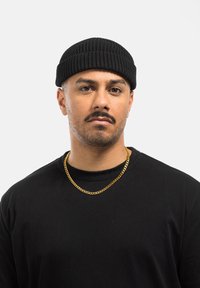 Sort ribbet beanie; rund form. Sort t-shirt med rund halsudskæring. Guld kæde halskæde; glat, sammenkoblet design. Neutral baggrund.