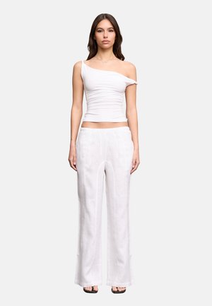 Donna in piedi che indossa un top bianco monospalla arricciato, pantaloni bianchi a gamba larga e sandali neri open toe su uno sfondo semplice.