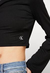 Mustassa ribbedoitu lyhythihainen paita, jossa on valkoinen brodeerattu "CK" -logo vasemmalla puolella, yhdistettynä vaaleansinisiin farkkuihin.