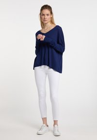 Maglietta a maniche lunghe blu navy con scollatura a V e vestibilità rilassata, abbinata a jeans skinny bianchi e sneakers bianche su uno sfondo neutro.