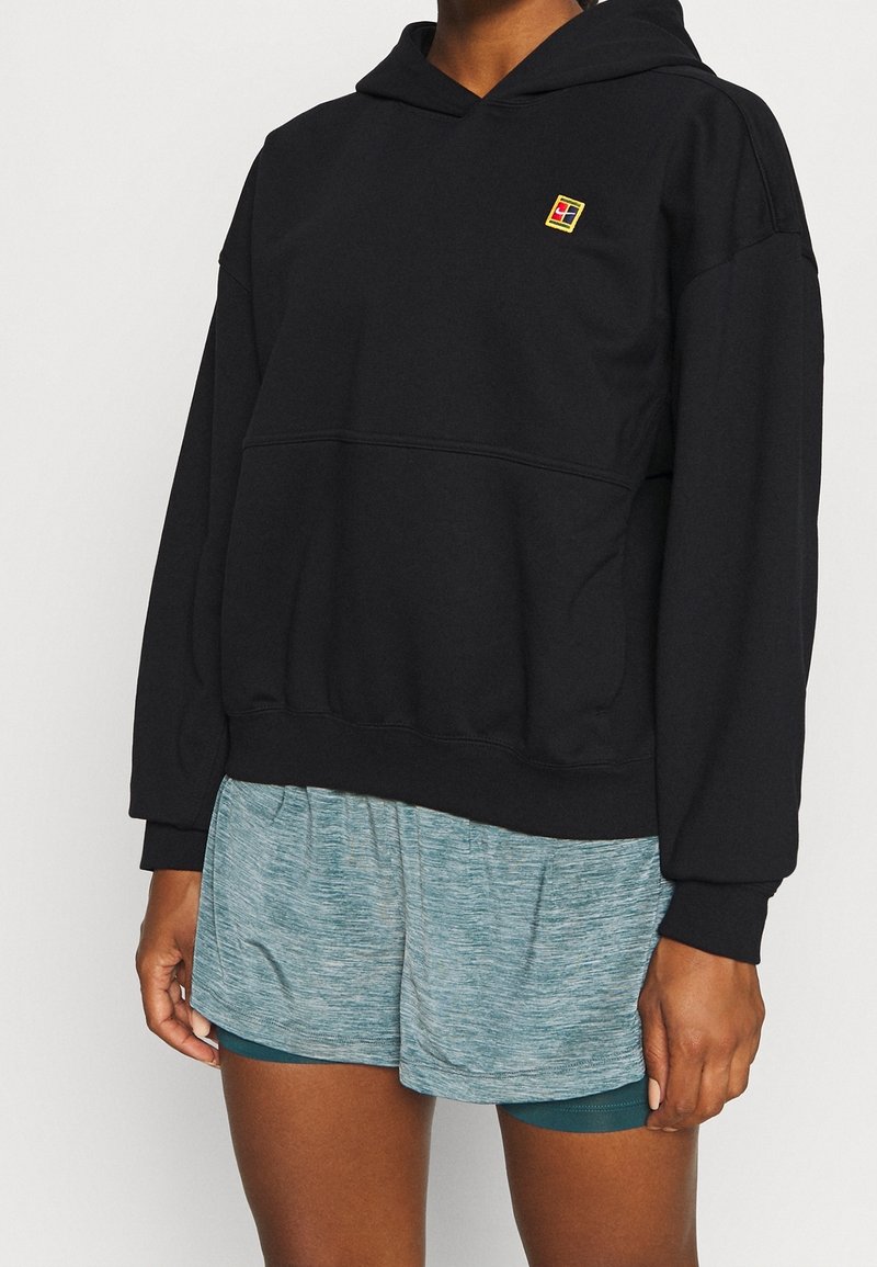 Personne portant un sweat à capuche noir avec un petit logo coloré sur la poitrine et un short de sport bleu clair.