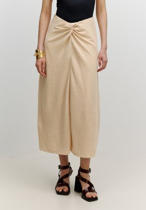 Beige rok tot de knie met een gedraaide voorkant. Gemaakt van een gestructureerde stof. Gepaard met donkerbruine sandalen met hak en bandjes.