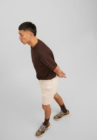 T-shirt marron à manches courtes, shorts beiges et baskets avec des accents orange et gris. Pose debout avec les mains jointes derrière.