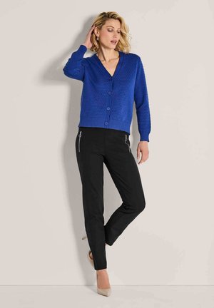 Vrouw staat met haar hand op haar hoofd, gekleed in een gestructureerde blauwe knoop cardigan, zwarte broeken met ritsen en beige hoge hakken.