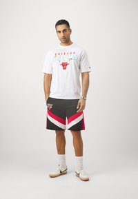 Bela majica s rdečim in teal grafičnim napisom "Chicago Bulls", črne hlače s rdečim in belim zigzag vzorcem, bele nogavice in bele superge.