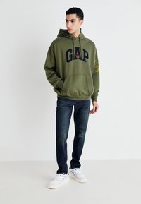 Olivgrön hoodie med marinblå "GAP"-logga, ficka fram och gul skrivdetalj på ärmen. Kombination med mörkblå jeans och vita sneakers.