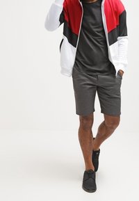 Tenue sportive pour homme comprenant une veste zippée rouge, blanche et noire, un t-shirt noir, un short gris et des baskets noires.
