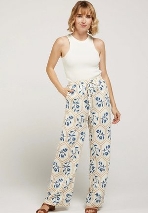 Femme portant un haut blanc sans manches et un pantalon large à motifs floraux bleus, debout avec une main dans la poche.